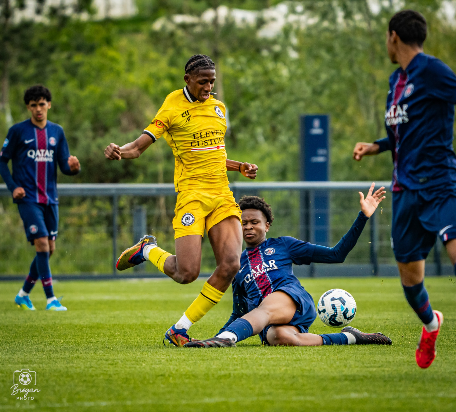 An AR Elite player in yellow competing for the ball against a Paris Saint-Germain defender.