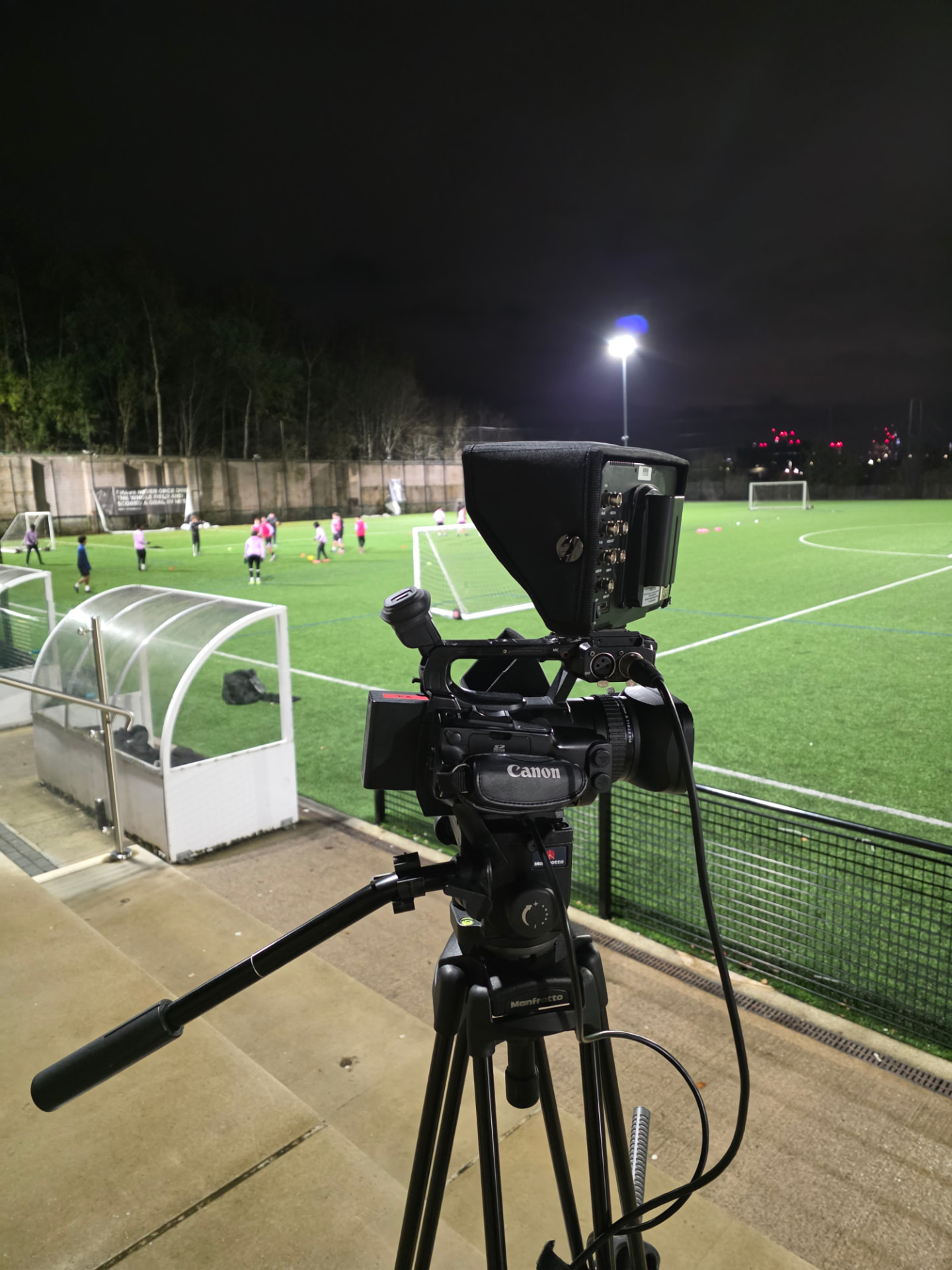 A professional camera on a tripod filming a floodlit evening football session.